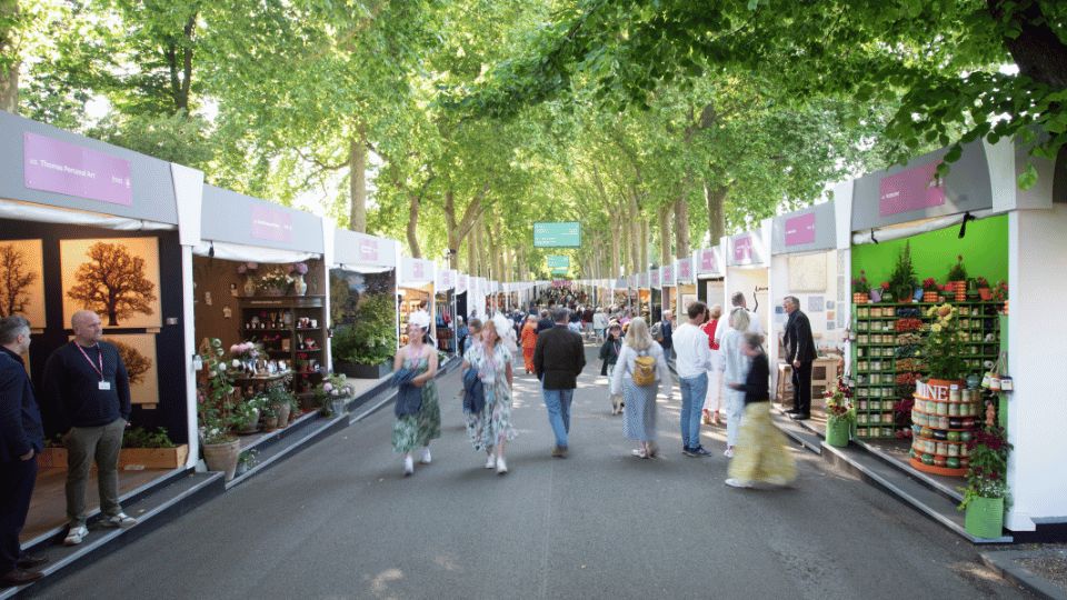 Visitors stroll along a tree-lined avenue at the RHS Chelsea Flower Show, browsing colourful exhibition stands filled with plants.